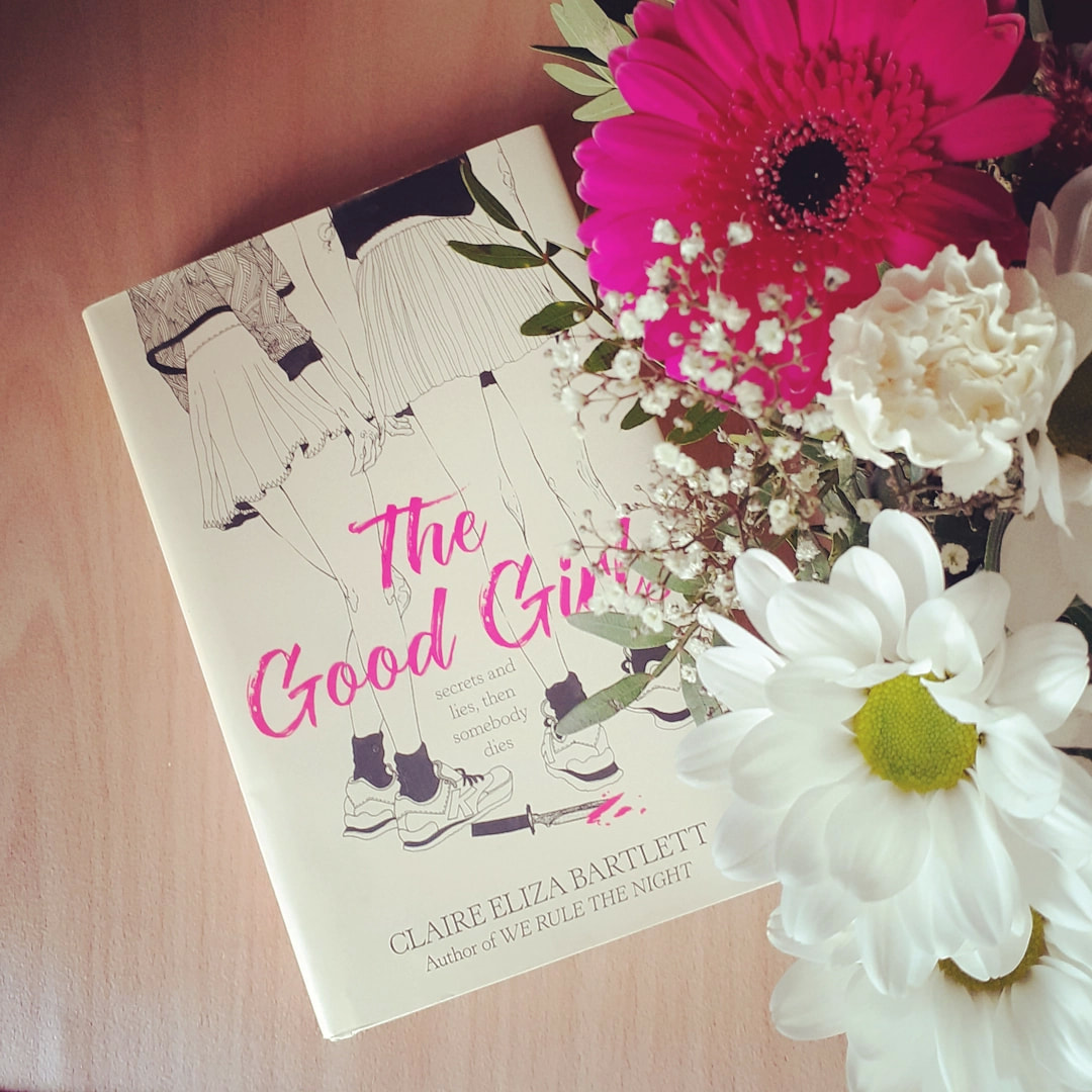 Photo du roman The Good Girls, de Claire Eliza Bartlett, posé sur un meuble en bois, avec des fleurs blanches et roses à côté